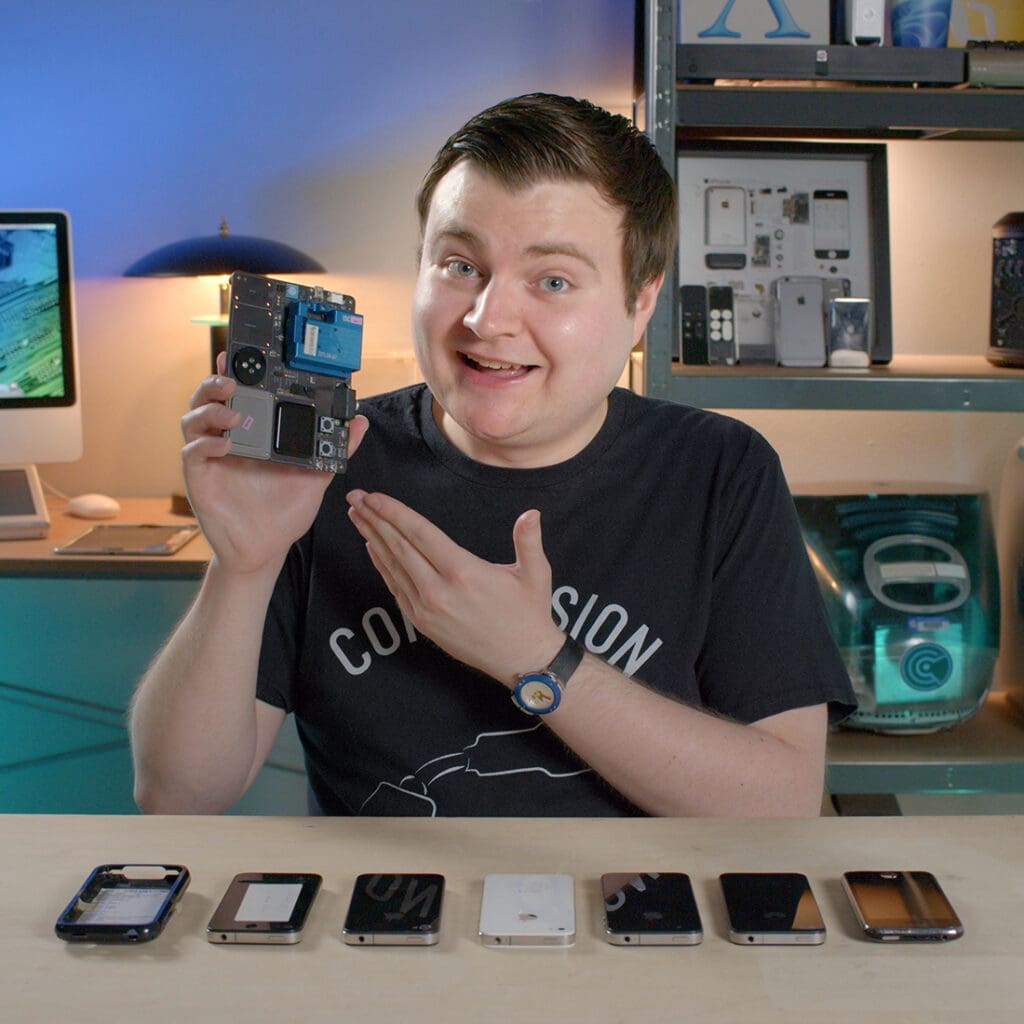 Krazy Ken is sitting in his lair while holding an Apple Watch breakout board. 5 iPhone 4 prototypes sit on the desk in front of him, as well as an iPhone stealth case and an iPhone 3Gs.