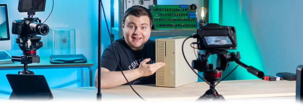 Behind-the-scenes picture of Krazy Ken in his studio, showing a camera in the foreground and behind his shoulder. He's smiling and gesturing toward a prototype Apple Macintosh Quadra 700 sitting on the desk.